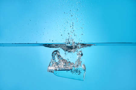 Bottle of cosmetic product in water with splashes on color background
