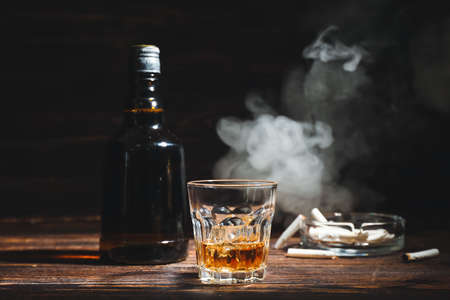 Bottle and glass of cold whiskey, cigarettes on wooden table against dark backgroundの写真素材
