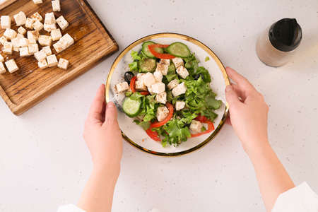 Woman with plate of fresh Greek salad on table in kitchenの写真素材