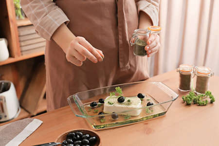 Woman adding spices into baking dish with tasty feta cheese on table in kitchen, closeupの写真素材