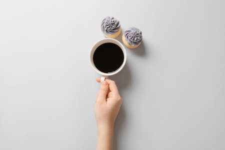 Female hand with cup of coffee and cupcakes on light background