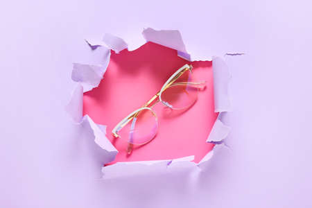 Stylish eyeglasses visible through torn paper on color backgroundの写真素材