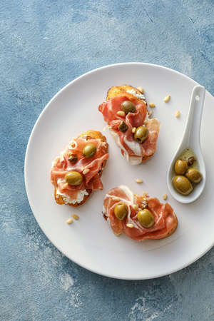 Tasty bruschettas with cheese, prosciutto and olives on color background