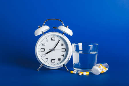 Alarm clock with glass of water and pills on color background