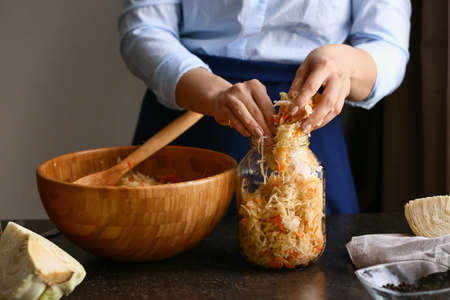 Woman putting tasty sauerkraut into glass jar on table in kitchen, closeupの写真素材