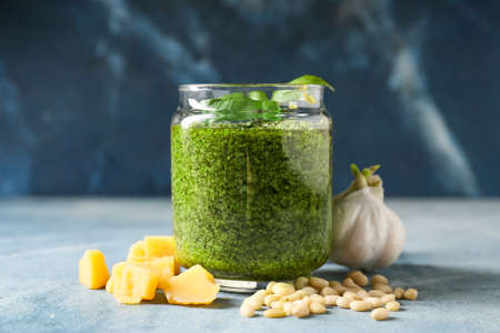 Glass jar with tasty pesto sauce, cheese, pine nuts and garlic on color background, closeupの写真素材