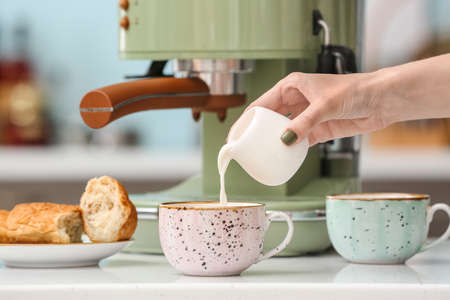 Woman pouring milk into cup of fresh coffee on table in kitchenの写真素材