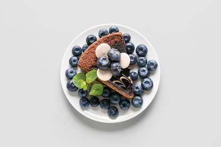 Plate with delicious piece of chocolate cake and blueberries on light background