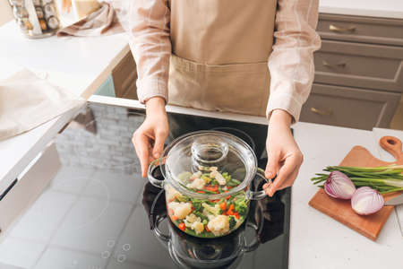 Woman with cooking pot of healthy vegetables on electric stove in kitchen