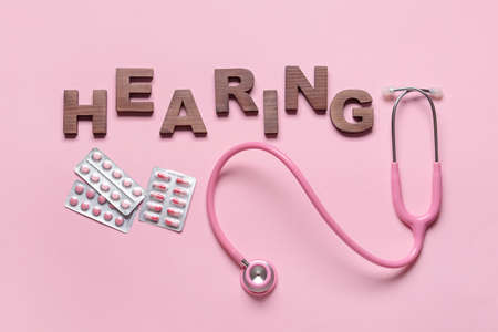 Word HEARING with stethoscope and pills on color background