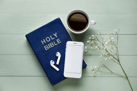 Holy Bible, earphones, mobile phone, gypsophila flowers and cup of coffee on color wooden background