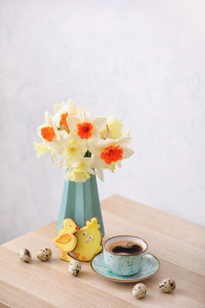 Vase with beautiful daffodils, quail eggs and cup of coffee on table against light background