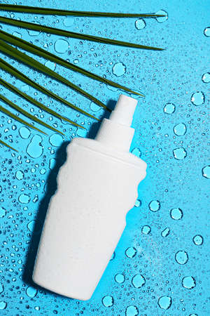 Bottle of sunscreen and palm leaf on color background with water drops