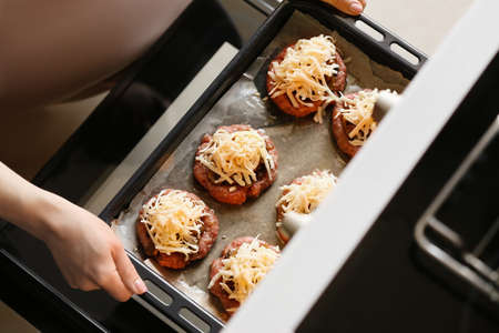Woman baking tray with minced meat boats, mushrooms and cheese into ovenの写真素材