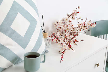 Beautiful blossoming branches, cup of tea, air freshener and pillow on chest of drawers in roomの写真素材