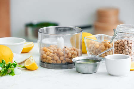 Blender with chickpeas and ingredients for hummus on table in kitchenの写真素材