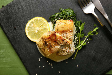 Board with tasty baked cod fillet, pesto sauce and lemon on color wooden background