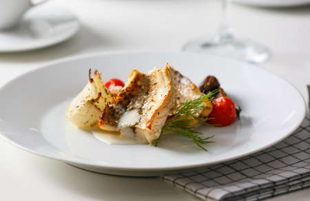 Plate with tasty baked cod fillet, vegetables and sauce on table in restaurant