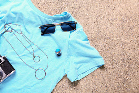 Stylish female accessories with t-shirt and photo camera on sand