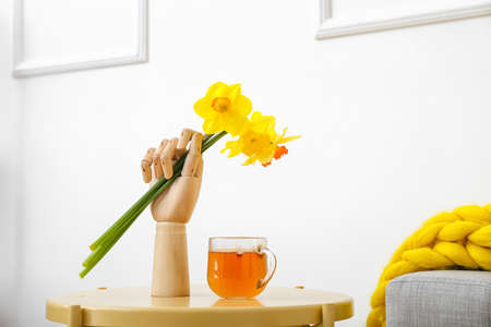 Mannequin hand with narcissus flowers and cup of tea on table in room