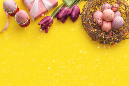 Nest with painted Easter eggs, gift and tulip flowers on yellow background with space for text
