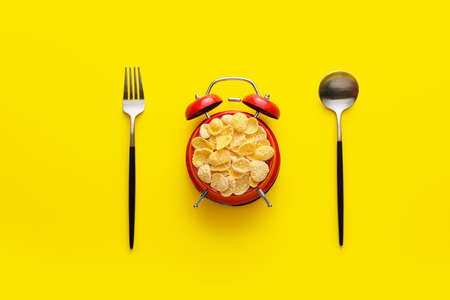 Alarm clock filled with flakes, spoon and fork on yellow backgroundの写真素材