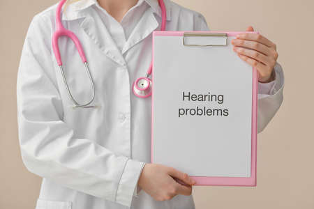 Female doctor holding clipboard with text HEARING PROBLEMS on color background