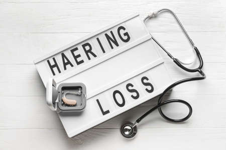 Board with text HEARING LOSS, aid and stethoscope on white wooden background
