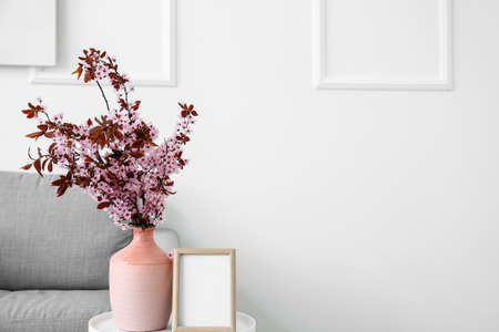 Vase with blossoming branches and blank frame on table near light wallの写真素材