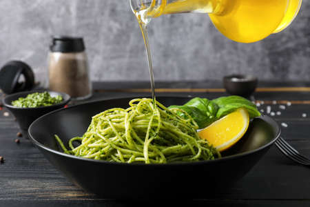 Pouring of olive oil from bottle onto plate with tasty pesto pasta on tableの写真素材