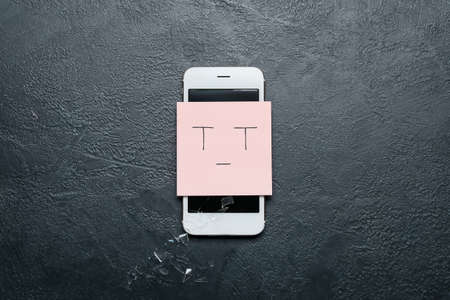 Damaged mobile phone with crying smile and scattered glass on dark backgroundの写真素材