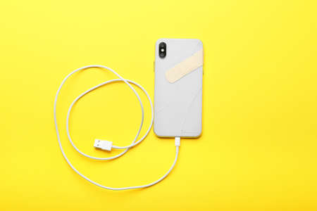Broken mobile phone with medical plaster and charging cord on yellow background