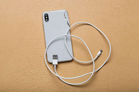 Damaged mobile phone and charging cord on color background