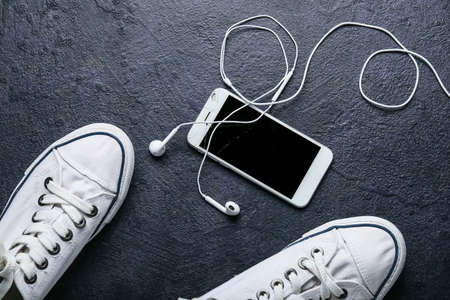 Mobile phone with broken screen, earphones and shoes on dark background