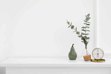 Vase with eucalyptus branches and alarm clock on shelf near light wall