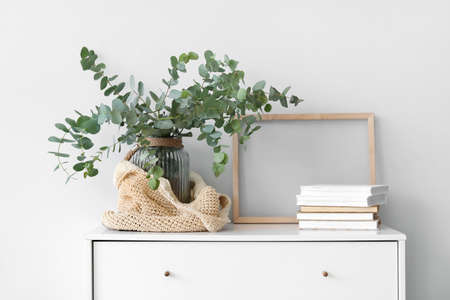 Vase with eucalyptus branches and books on chest of drawers near light wall