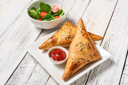 Plate with tasty Uzbek samsa and tomato sauce on white wooden background