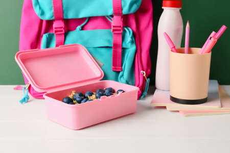 Lunch box with berries, bottle of drink, backpack and stationery on table in classroom