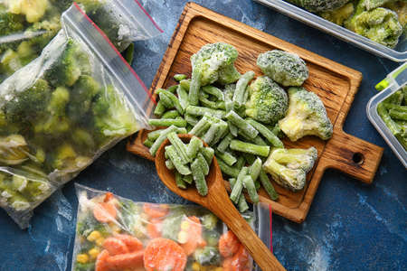 Wooden board and plastic bags with different frozen vegetables on color background