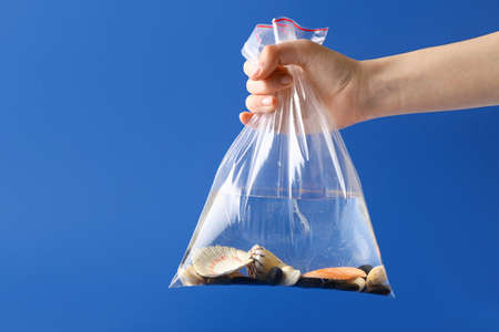 Human hand with water and sea shells in plastic bag on color background. Ecology concept