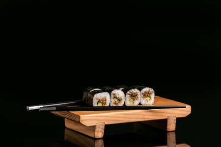 Wooden board with delicious sushi rolls and chopsticks on dark background
