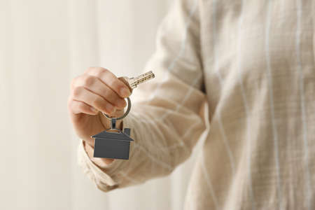 Woman holding key from house with keychain on light background