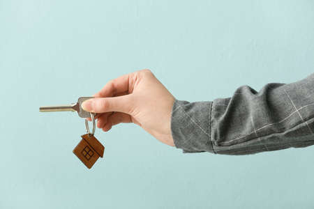 Real estate agent holding key with house shaped keychain on color background