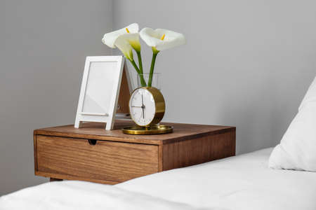 Alarm clock with calla lilies and frame on wooden bedside table in bedroomの写真素材