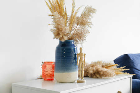 Vase with bouquet of dried flowers and wooden mannequin doll on chest of drawers near light wallの写真素材