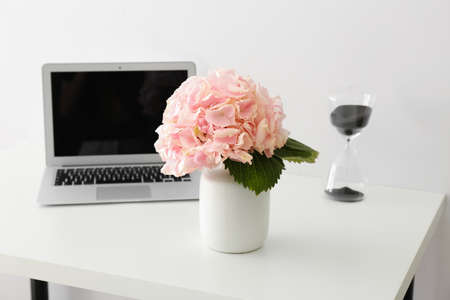 Vase with beautiful hydrangea flowers, modern laptop and hourglass on tableの写真素材