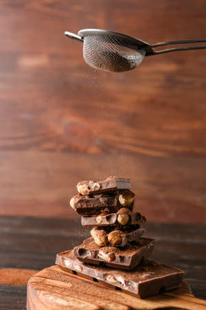 Sprinkling of cacao powder onto pieces of tasty chocolate with nuts on wooden background