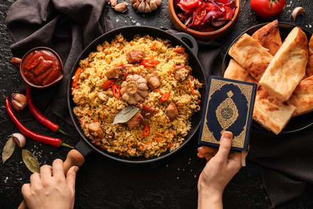 Female hands with frying pan of tasty Asian pilaf and Quran on dark background