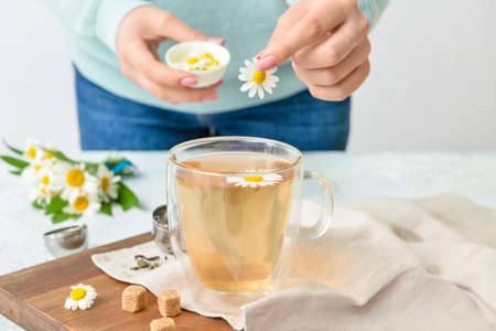 Woman putting chamomile flower into cup with tea on table