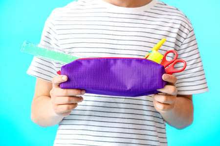 Little boy holding pencil case with stationery on color background